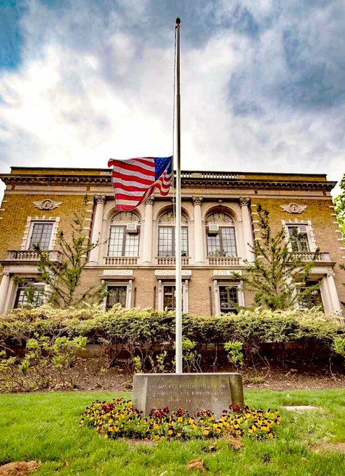 A stately brick building with classical columns stands under a cloudy sky. An American flag is at half-mast on a white flagpole in front of the building. A circular arrangement of yellow and purple flowers surrounds the base of the flagpole, set upon a well-maintained grassy lawn.