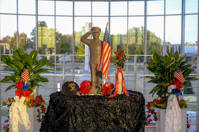 A statue of a saluting soldier stands on a table. There are plants and flags on each side of the table.