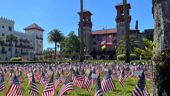 A grassy area filled with numerous small American flags in front of the historic Lightner Museum in St. Augustine, Florida. The building features Spanish-style architecture with arches and palm trees nearby. Two larger flags are mounted near the entrance.