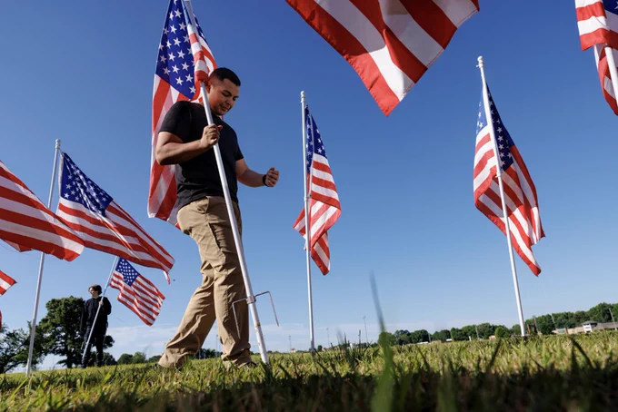 A person places an American flag into the ground in a grassy field filled with waving U.S. flags under a bright blue sky.