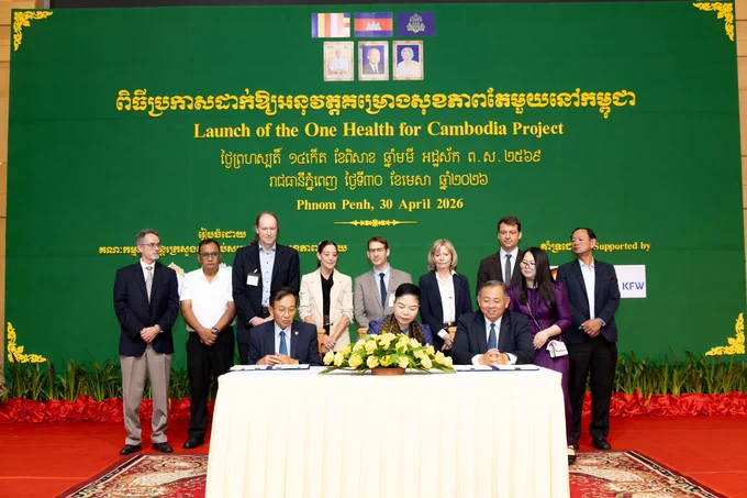 A group of individuals stands on a stage in front of a green backdrop with yellow text, including the phrase "Launch of the One Health for Cambodia Project." Two people are seated at a table signing documents, while others stand behind them. Signage indicates this event is supported by various organizations, with Phnom Penh as the location and the date listed as April 30, 2026.