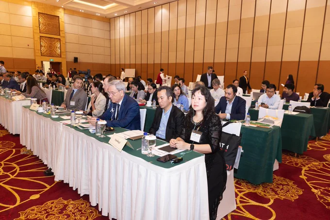 A large conference room is filled with attendees seated at tables covered with green and white cloths. The attendees are focused on a presentation or speaker. The room has high ceilings and warm lighting. In the background, more people are engaged in discussion.