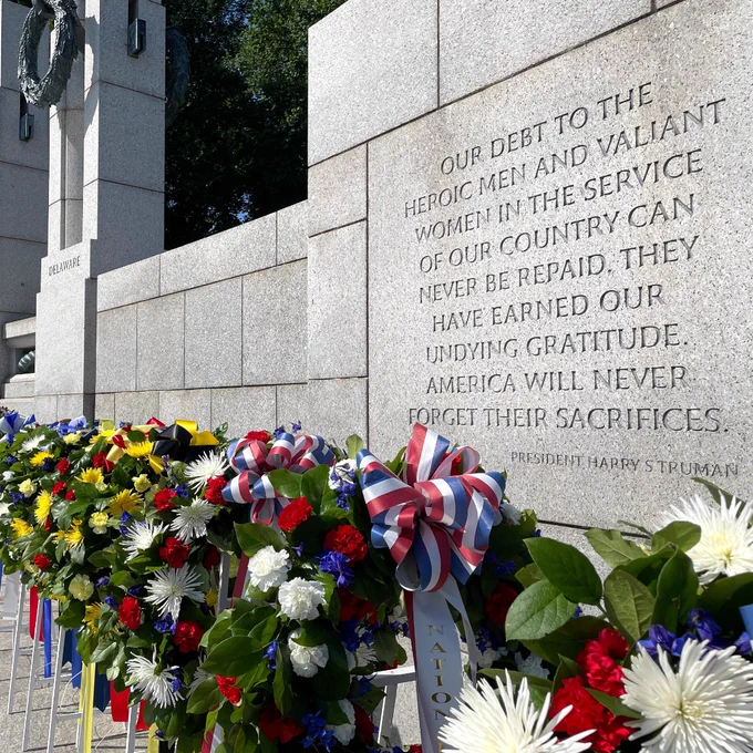 A line of colorful flower wreaths on easels standing in an outdoor military memorial in front of a gray stone wall etched with the quote: OUR DEBT TO THE HEROIC MEN AND VALIANT WOMEN IN THE SERVICE OF OUR COUNTRY CAN NEVER BE REPAID. THEY HAVE EARNED OUR UNDYING GRATITUDE. AMERICA WILL NEVER FORGET THEIR SACRIFICES.