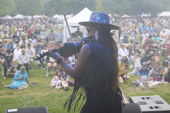 Female violinist wearing a wide-brimmed hat and sunglasses performing on stage at an outdoor festival with a large seated audience.
