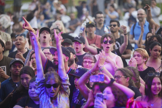 Crowd of people at an outdoor event raising hands and cheering with colorful lights illuminating the scene.