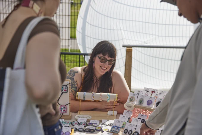 Vendor showcasing handmade jewelry including bracelets and earrings at an outdoor market stall with customers browsing.