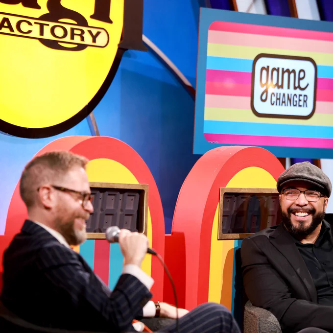 Two men are seated on a colorful set, engaged in conversation, with vibrant graphics and a "Game Changer" sign in the background.