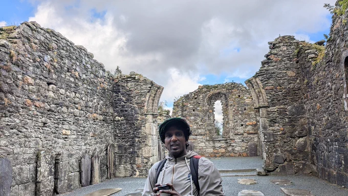 [Image: Wearing a dark cap and light brown jacket, Hari Srinivasan stands inside stone castle ruins along the coast of Northern Ireland.]