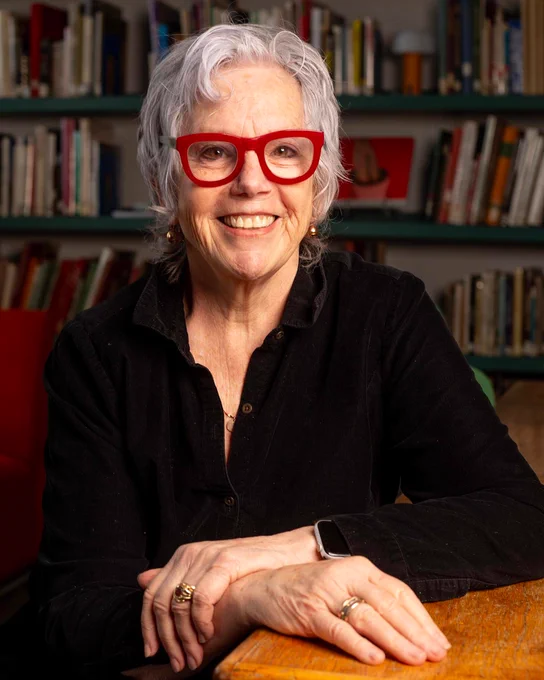 A woman with short gray wavy hair and bright red-framed glasses smiles warmly at the camera while seated at a wooden table. She wears a black button-up shirt and has her hands folded on the table, displaying several rings and a smartwatch on her wrist. Behind her, a bookshelf is filled with colorful books and a red container, suggesting an office or study setting.