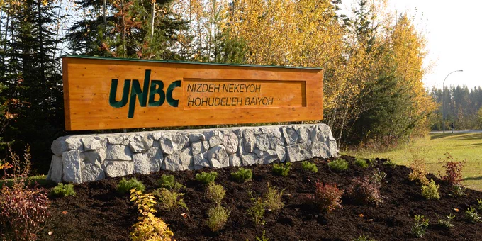 A wooden sign with the text "UNBC" and additional writing in another language, situated on a stone base. The sign is surrounded by landscaped greenery and backed by autumn trees.