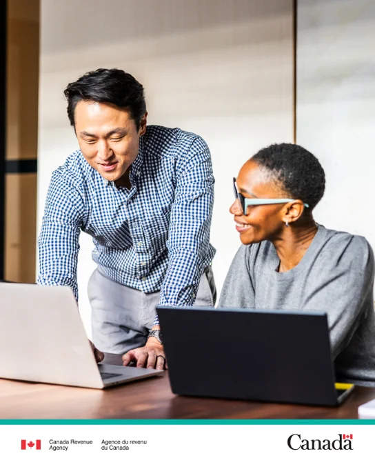 Two people with open laptops, one is standing and one is sitting as they both look towards one of the laptop screens.