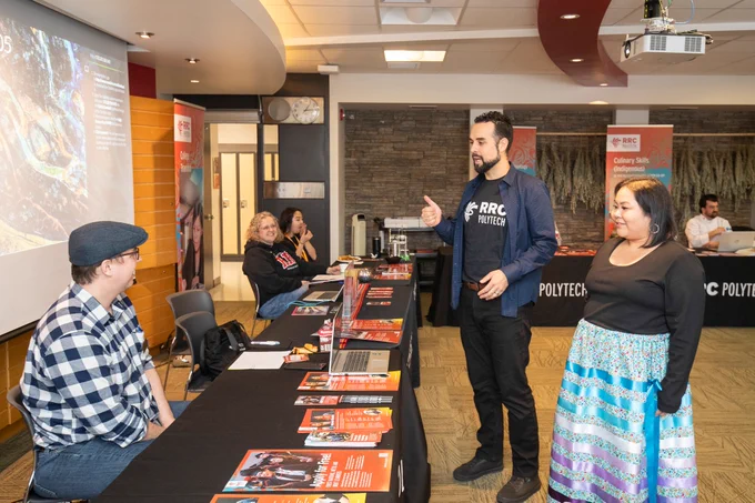 Alt Text: Staff and prospective students speak at an RRC Polytech information event, with program brochures and displays set up on tables throughout the room. Indigenous program banners and a presentation screen are visible in the background as attendees connect and ask questions.