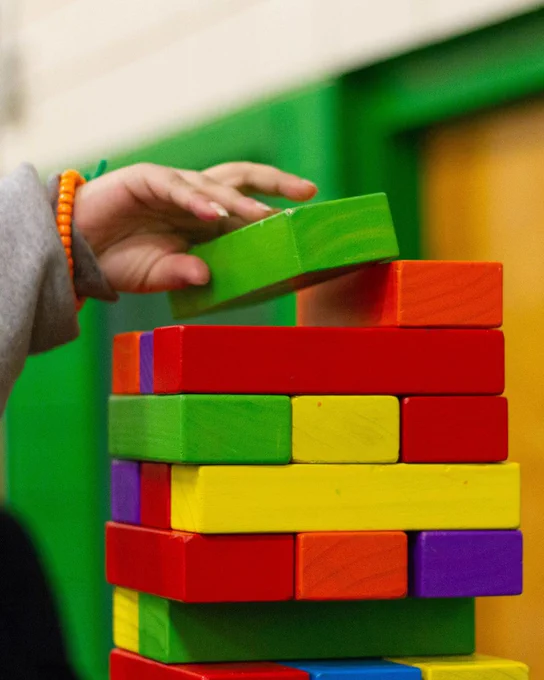 Photograph shows a child's hands putting a colourful wooden block on top of a tower of colourful wooden blocks. 