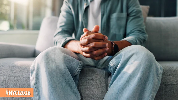 Person sitting on a gray couch with hands clasped, wearing a denim shirt and jeans in a softly lit room.
