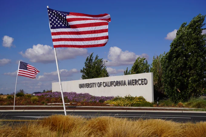 American flags waving near the University of California Merced entrance sign under a clear blue sky.