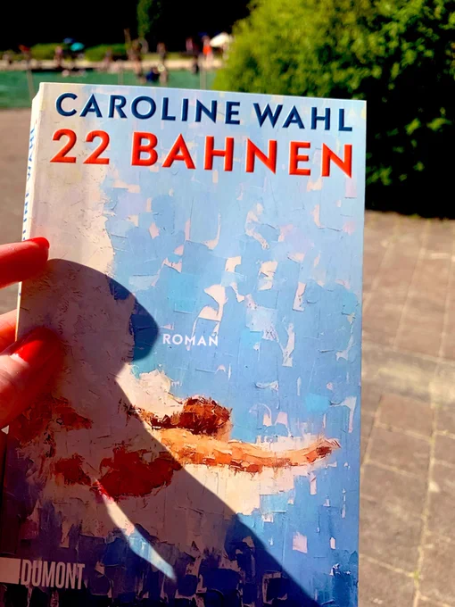 Ein Foto aus dem Freibad. Im Hintergrund sieht man das Becken. Im Vordergrund halte ich das Buch „22 Bahnen“ von Caro Wahl.