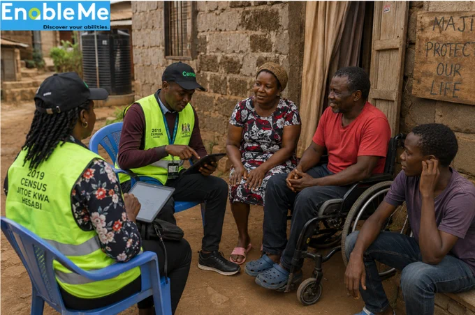 Kenya national census officers conduct a household interview with community members, including a wheelchair user, using tablets in a mixed rural-urban neighborhood.