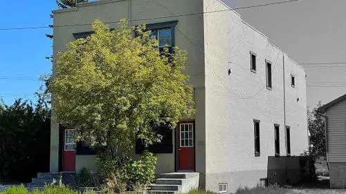 A two-story beige brick building with red doors and black-trimmed windows is partially shaded by a lush green tree. A neatly maintained lawn surrounds the structure.