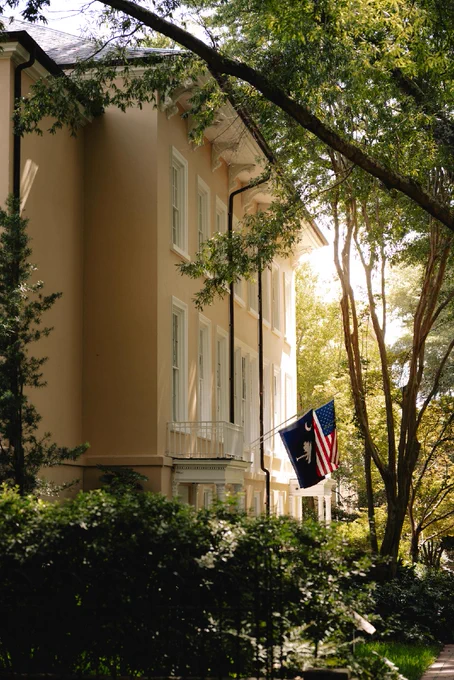 The USC President's house with white-framed windows is partially visible through lush green trees. Two flags hang on the building, one being the United States flag and the other a South Carolina flag. Sunlight filters through the foliage, casting soft light on the scene.