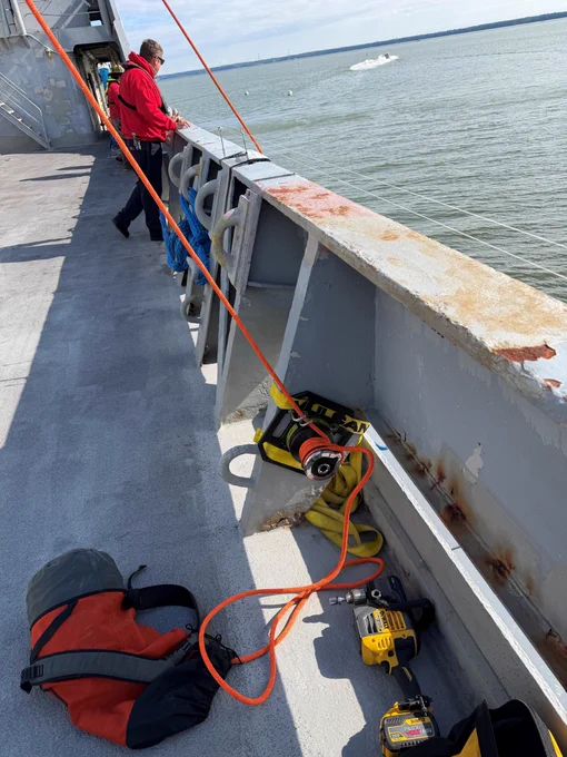 A person in a red jacket stands on the deck of a ship, gazing at the water. A rope is attached to the railing, connected to a winch and extending over the side. A drill and backpack lie on the deck. A distant boat is visible on the water.