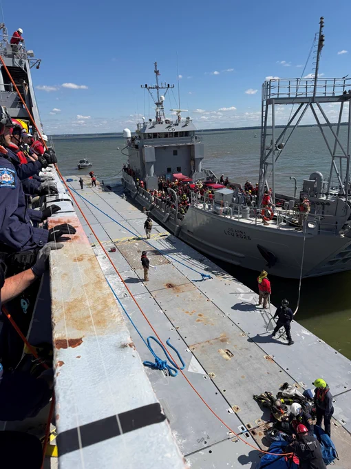 A Coast Guard vessel is docked alongside a larger ship, with several crew members visible on both vessels. Some individuals are gathered near rescue equipment on the deck. The waterway is calm, and the sky is clear with a few clouds.