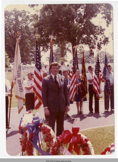 Man in suit standing solemnly in front of wreaths with multiple American flags and color guard behind him at a memorial event.