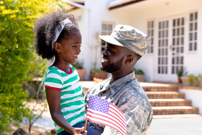 A smiling person in a military uniform holds a child who is holding an American flag. They are standing outside a house, surrounded by greenery, and both appear happy.