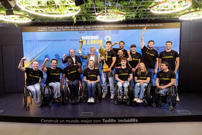 Grupo de personas en un escenario, varias en silla de ruedas, posan juntas con camisetas del equipo de baloncesto en silla de ruedas CD ILUNION junto al CEO de ILUNION, Alejandro Oñoro, que están levantando la copa de campeones de Europa. 