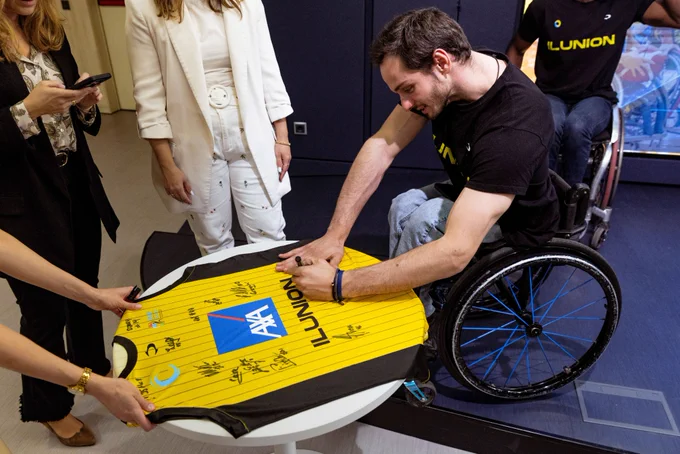 Persona en silla de ruedas firma una camiseta amarilla del CD ILUNION extendida sobre una mesa, rodeada de otras personas. La camiseta contiene varias firmas visibles.