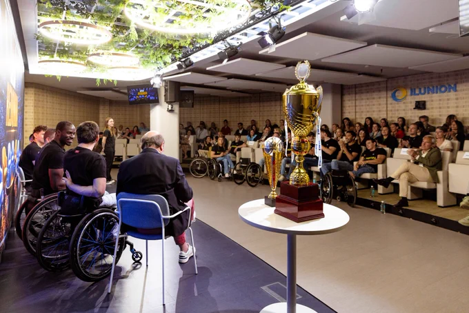 Vista de un evento con público sentado frente a un escenario. En primer plano, trofeos de campeones de europa de baloncesto en silla de ruedas sobre una mesa. En el escenario, varias personas, algunas en silla de ruedas, participan en una charla. Logotipo de ILUNION visible en la pared.