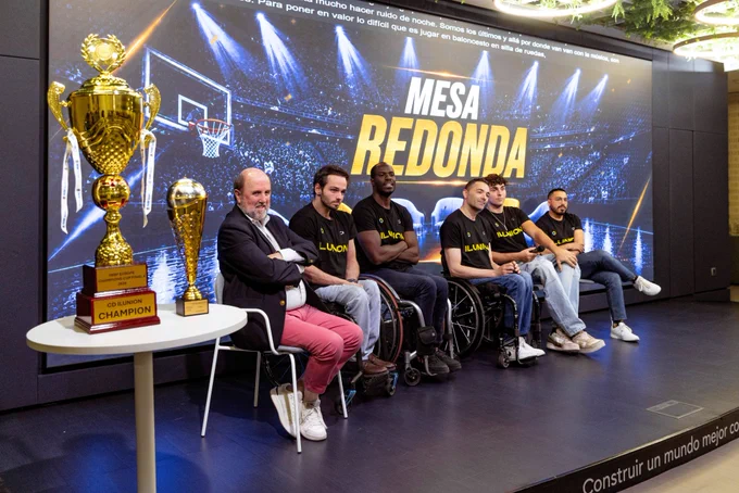 Personas sentadas en sillas y sillas de ruedas participan en una mesa redonda. A la izquierda, varios trofeos sobre una mesa. En la pantalla de fondo se lee “MESA REDONDA” con imagen de una cancha de baloncesto.
