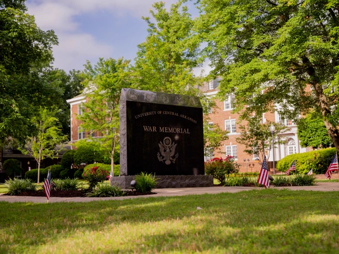 A serene outdoor scene featuring the University of Central Arkansas War Memorial, a large stone monument with engraved text. It is surrounded by a neatly maintained lawn, landscaping, and several small American flags. In the background, red brick buildings and lush green trees are visible under a partly cloudy sky.