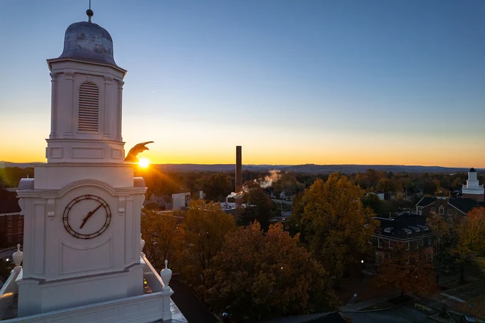 Aerial view of a clock tower at sunrise with smoke rising from a chimney in the background, surrounded by autumn trees and buildings.