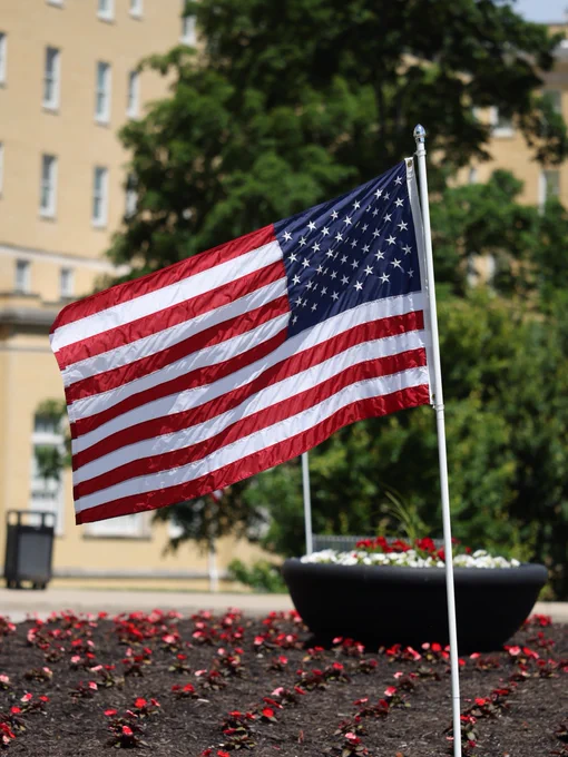 A United States flag waves in front of French Lick Springs Hotel, surrounded by a garden bed with small red and pink flowers. A large planter with flowers is visible in the background.