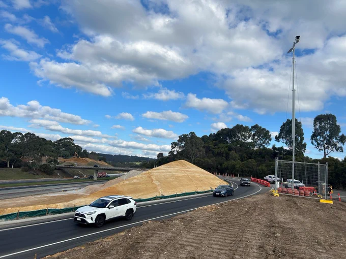 A road curves around a large mound of earth under a blue, partly cloudy sky. Cars move along the road, including a white SUV. In the background, there is an overpass and dense trees.