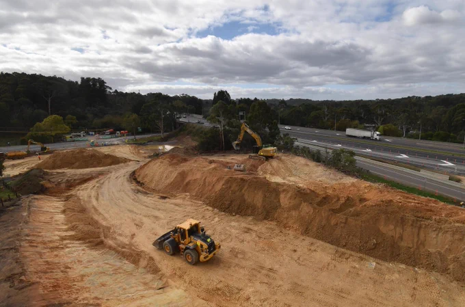 An aerial shot shows a construction site with heavy machinery like excavators and loaders, with a highway in the background carrying cars and a truck, set against a backdrop of trees and a cloudy sky.
