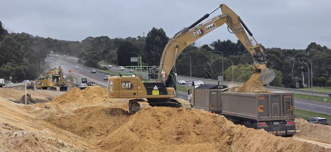 Excavator, branded 'CAT leed', filling sand into a truck on a highway construction site. Other construction vehicles are also present. Trees and an overcast sky are visible in the background. 'EX107@1', '340@1', 'YS 44FS@1'.