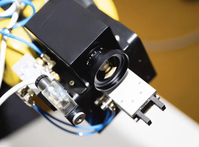 Close-up of an industrial camera sensor with lens mounted on a robotic system, connected by cables and components, used for machine vision or automated inspection in a controlled environment.