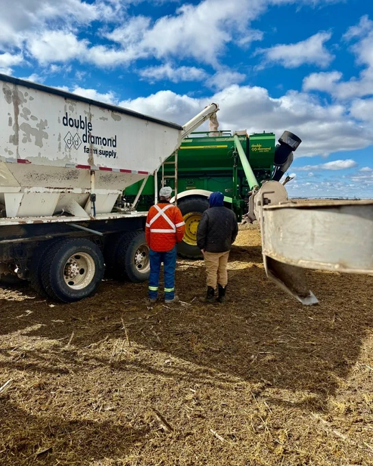 Two individuals stand next to a Double Diamond farm supply truck as it loads seed into a green air cart for seeding. The setting is a bare field under a partly cloudy sky.