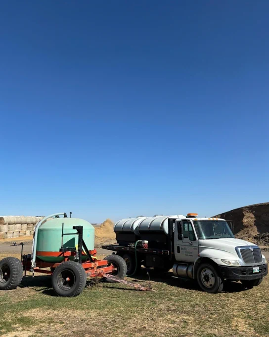 A white industrial truck with a large cylindrical tank attached, parked on grass under a clear blue sky. Hay bales are stacked in the background.