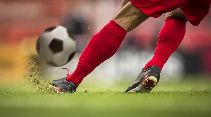 Close-up action shot of a soccer player's lower legs in red socks and black cleats kicking a black-and-white soccer ball on a green grass field, with bits of grass and dirt kicking up into the air.