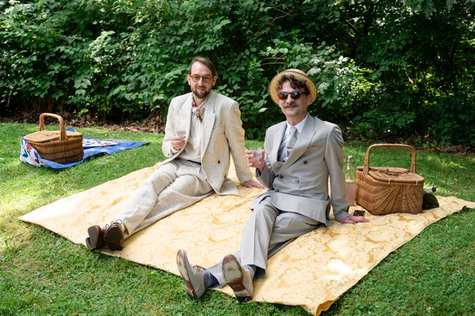 Two men in vintage suits relaxing on a blanket in a lush green park, each holding a drink with picnic baskets nearby.