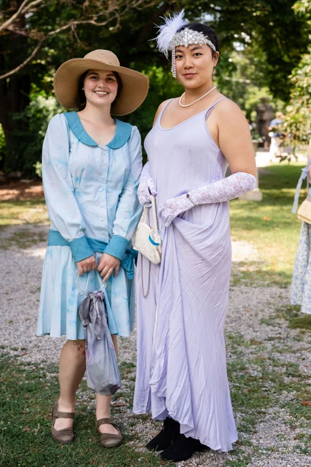 Two women dressed in vintage-style outfits stand outdoors on a gravel path surrounded by greenery.