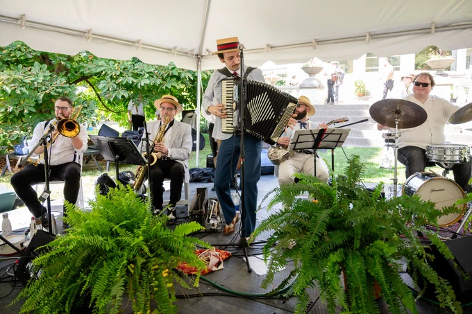 A five-piece band performs under a white tent, featuring accordion, trombone, saxophone, guitar, and drums surrounded by green plants.