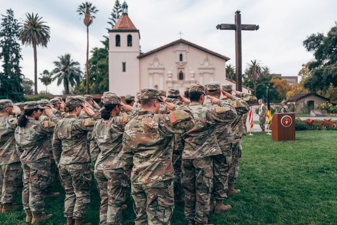 A group of uniformed individuals stand and salute outdoors in front of the historic Mission Santa Clara de Asís, with a large wooden cross and a podium nearby. Palm trees and other greenery are in the background.