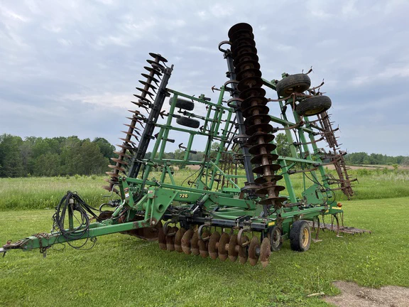 A large John Deere agricultural implement with multiple rotating discs is positioned on a grassy field. The equipment appears used for tilling soil, with both the machinery and sky in the background suggesting an outdoor rural setting.