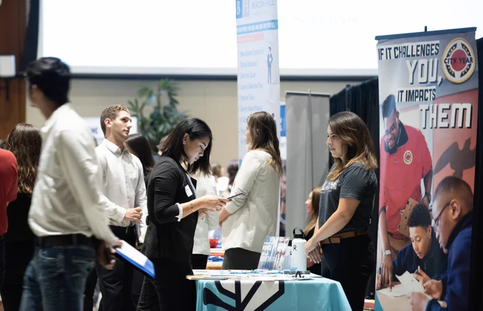 A busy job fair with several people interacting at different booths. One table displays a banner with "City Year" and an inspirational message. Attendees are exchanging information, and various promotional materials are visible on the tables.