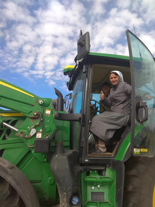 A Religious Sister of the Missionary Benedictine Sisters is seated at the controls of a large green tractor and holding open the glass tractor cab door with her left hand. She is looking at the camera and giving a "thumbs up" with her right hand. She is wearing a gray habit with gray jacket, white veil, gray and brown striped socks, and black shoes. A young man is next to her in the cab and smiling at the camera from over the Sister's shoulder.
