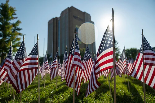 A field of American flags is displayed on a grassy area with the Kent State University Library in the background. The sun is shining brightly, casting shadows and creating a lens flare effect.