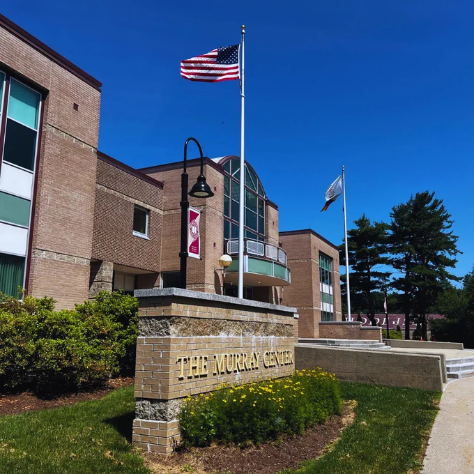 The image shows a building with a sign in front reading "The Murray Center." An American flag is flying on a flagpole at Rhode Island College. The building is brick with large windows, and there are trees and shrubs in the landscaping. The sky is clear and blue.